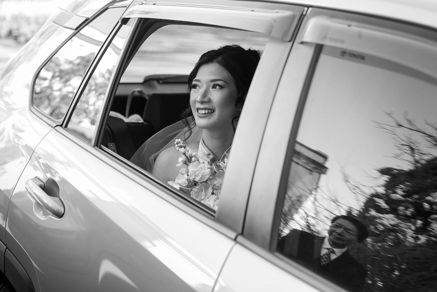 Black and white photo of a Bride smiling from the back seat window of a car with a reflection of her Dad in the window at St. Matthews.