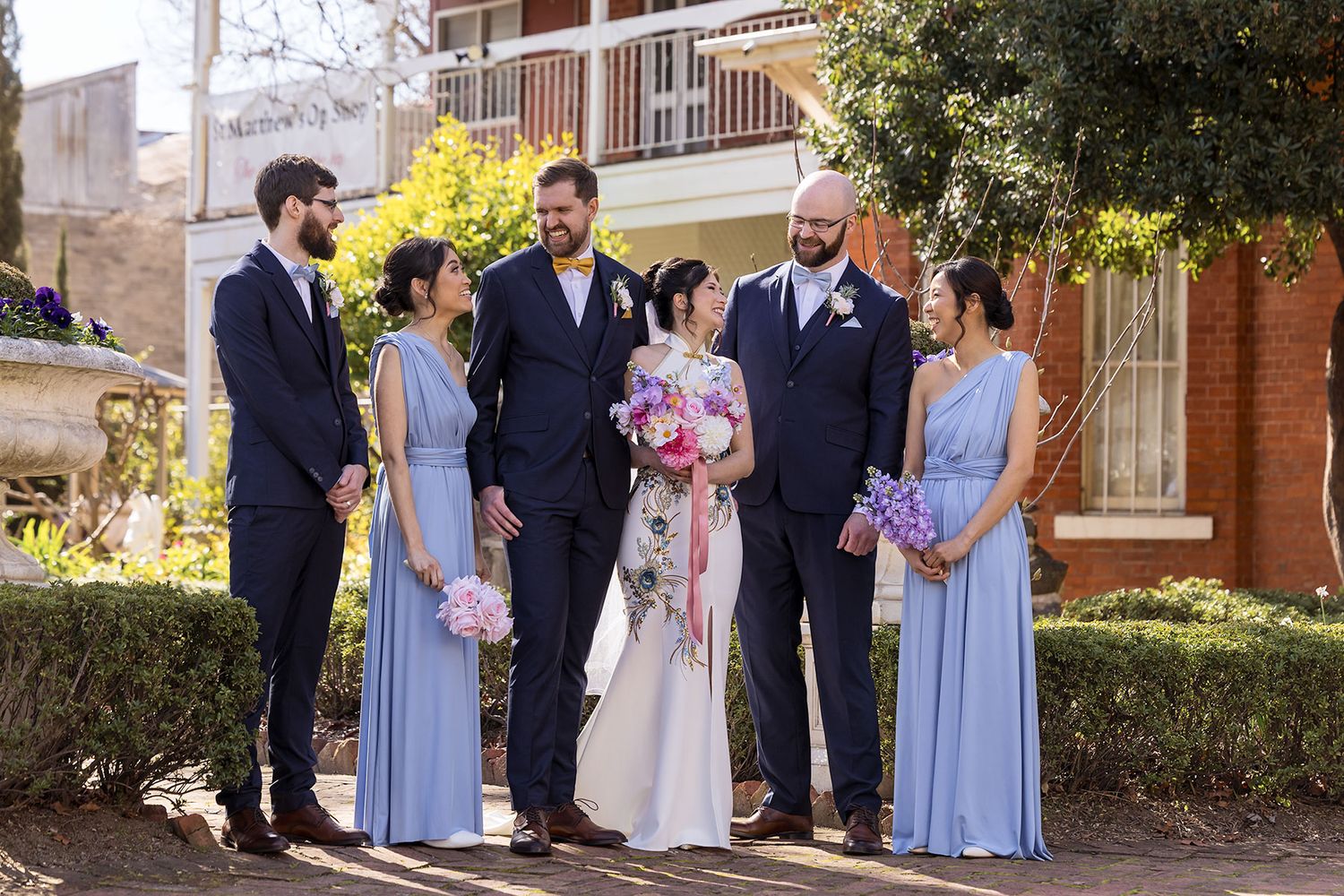Wedding party members in light blue and formal attire pose together outside a brick building in natural lighting.