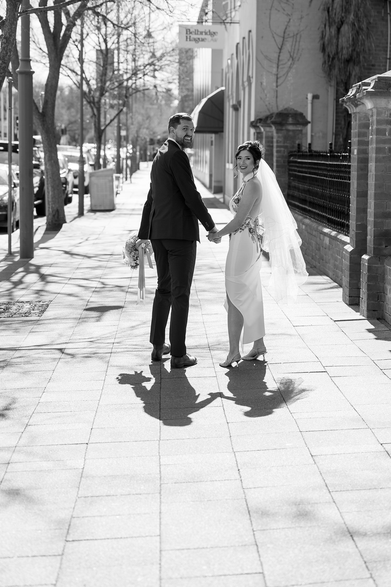 A black and white photograph of a bride and groom holding hands walking together on a sunny sidewalk in front of St. Matthews Church.