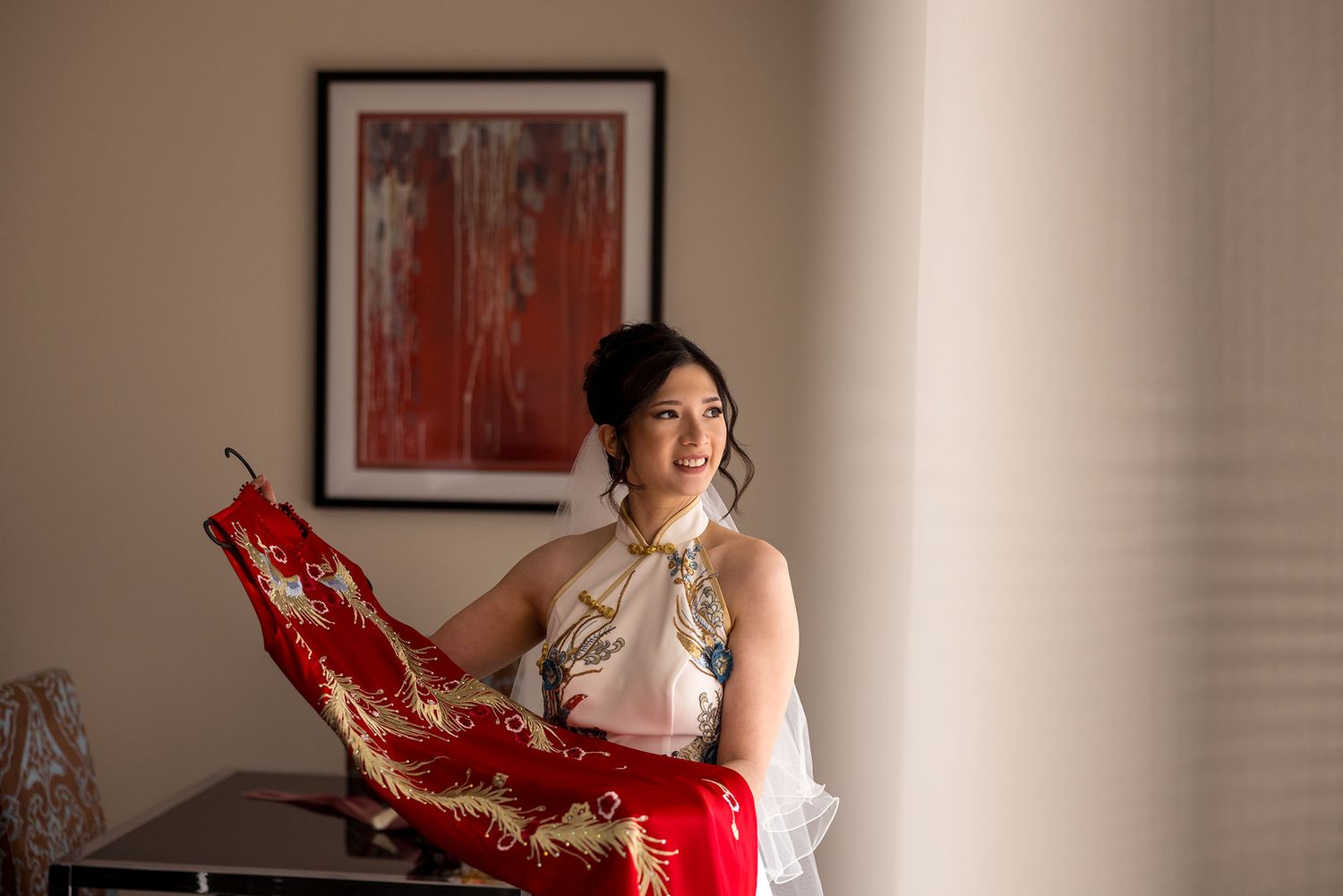 A Bride in traditional Chinese dress holding red qipao dress while posing indoors beside framed artwork at De Bortoli Rutherglen Estate.