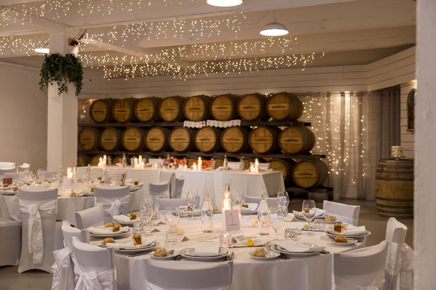 Elegant wedding reception venue with white tables, string lights, and rustic wine barrels displayed on the wall at De Bortoli Rutherglen.