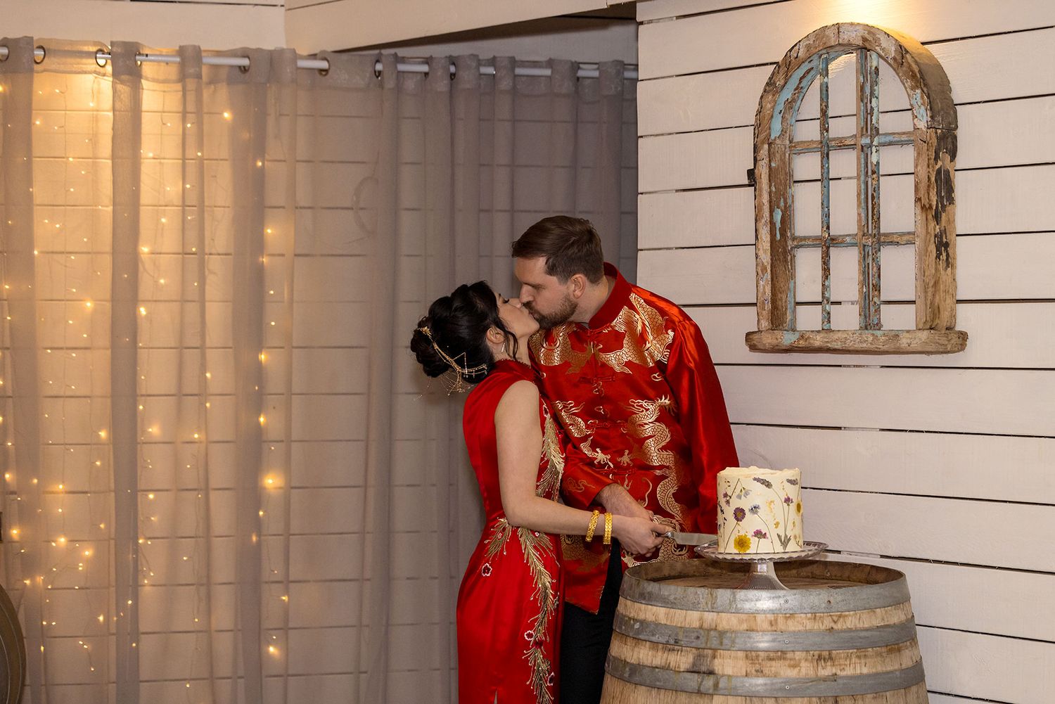 A romantic Bride and Groom in red clothing embrace near a rustic wine barrel with string lights creating a cozy atmosphere cut their cake.