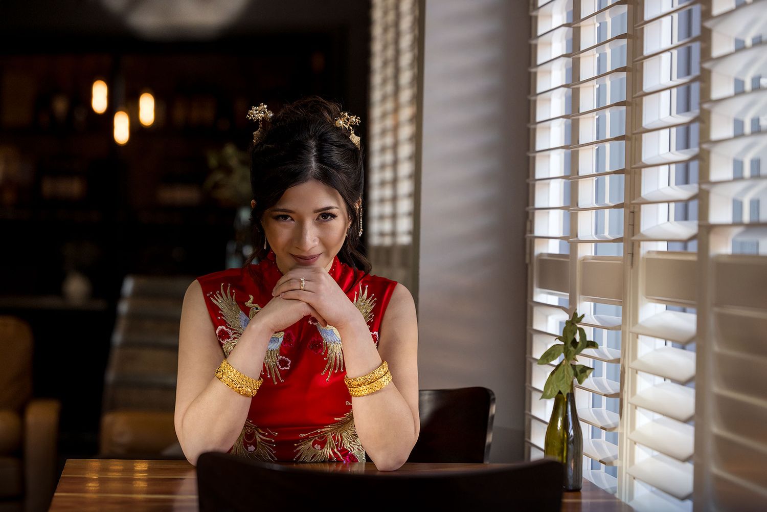 A Bride in a traditional red cheongsam dress sits thoughtfully at a table by window blinds in a dimly lit restaurant at DeBortoli Rutherglen