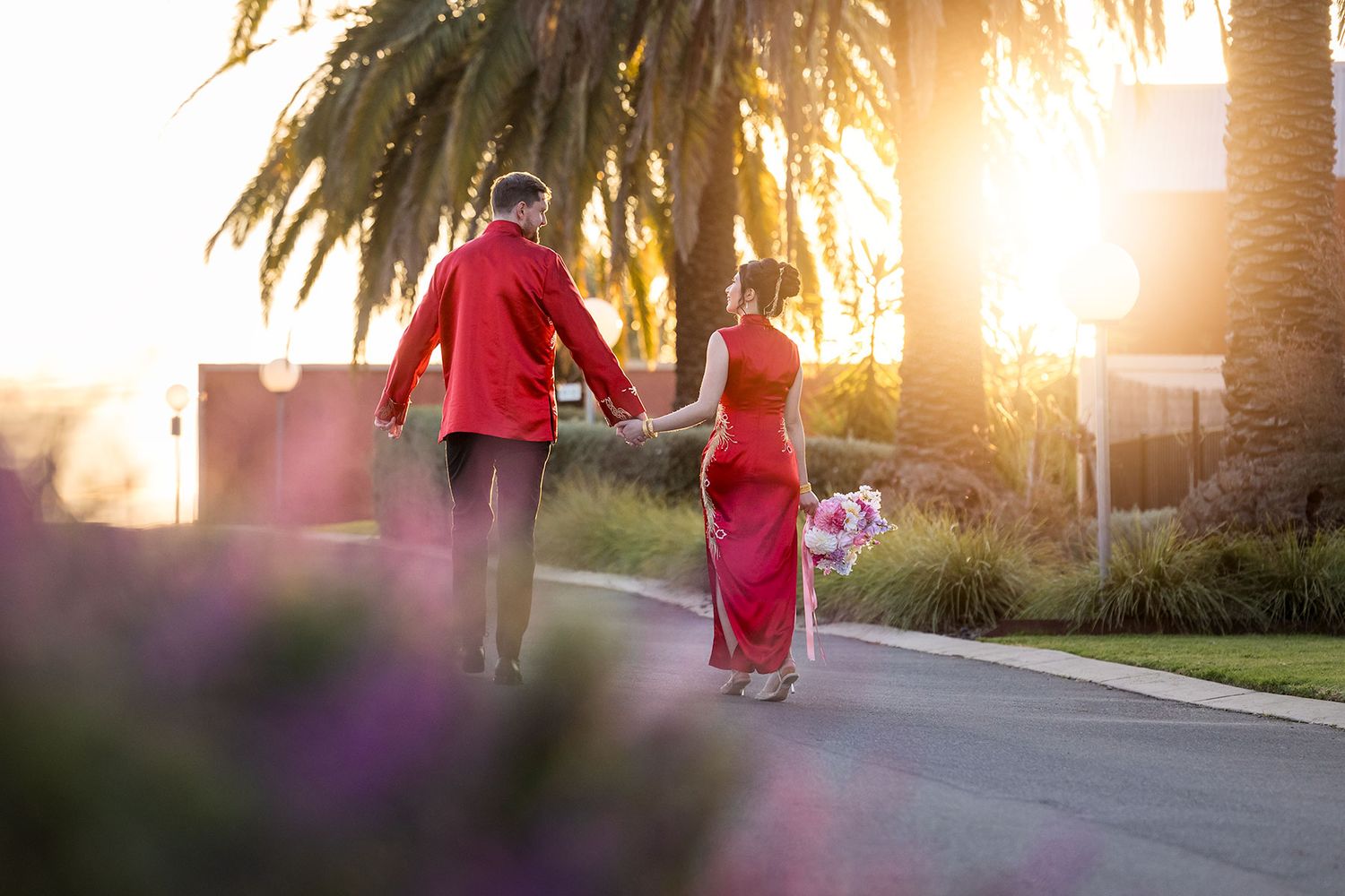 A Bride and Groom in traditional Chinese red outfits walk hand in hand down a palm tree-lined street at sunset at De Bortoli Rutherglen.