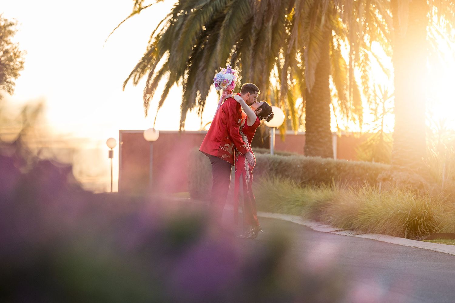 A Bride and Groom in traditional Chinese red outfits share a kiss on a palm tree-lined street at sunset at De Bortoli Rutherglen Estate.