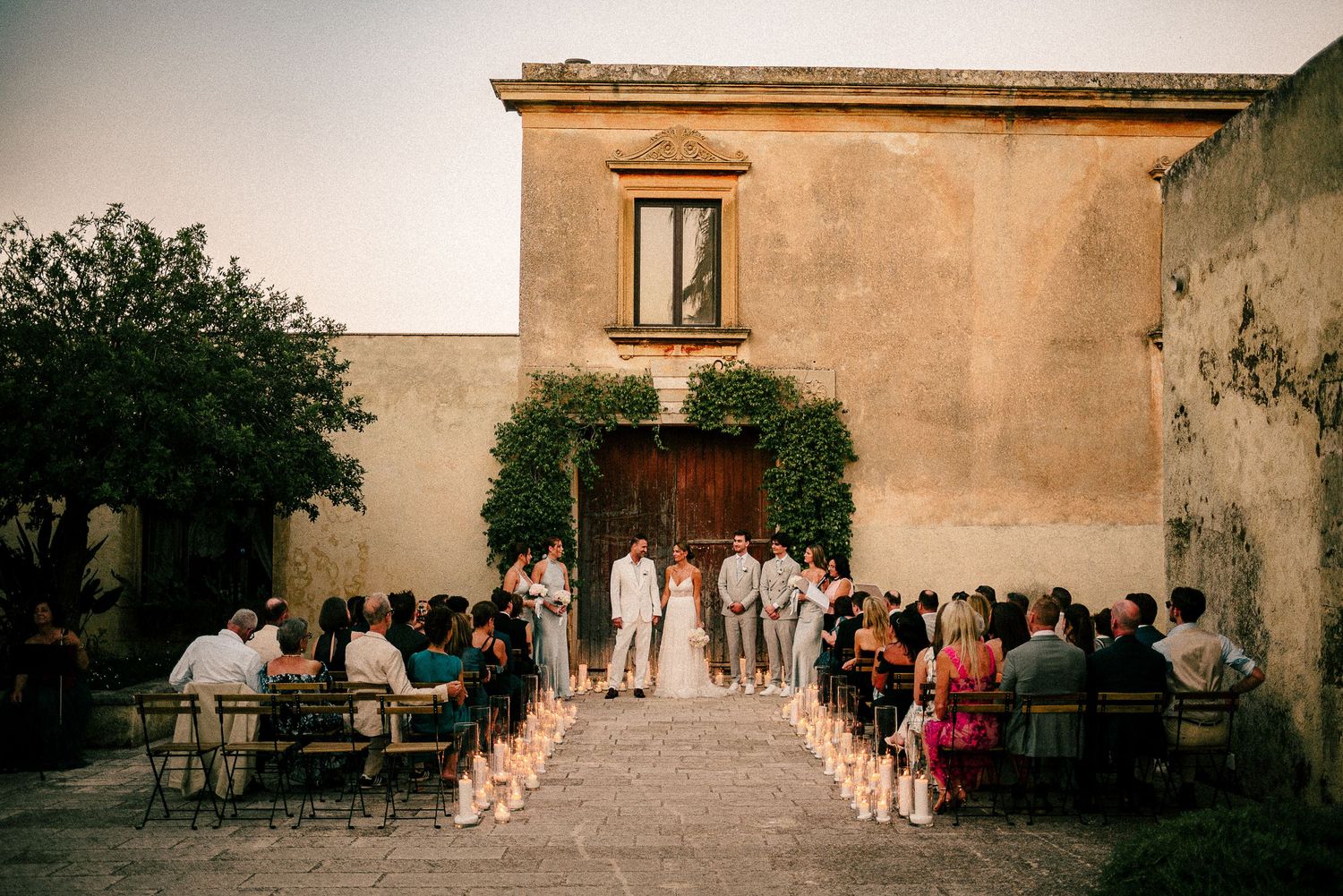 A romantic outdoor wedding ceremony takes place at dusk in front of an old stone villa with pathway lined by candles.