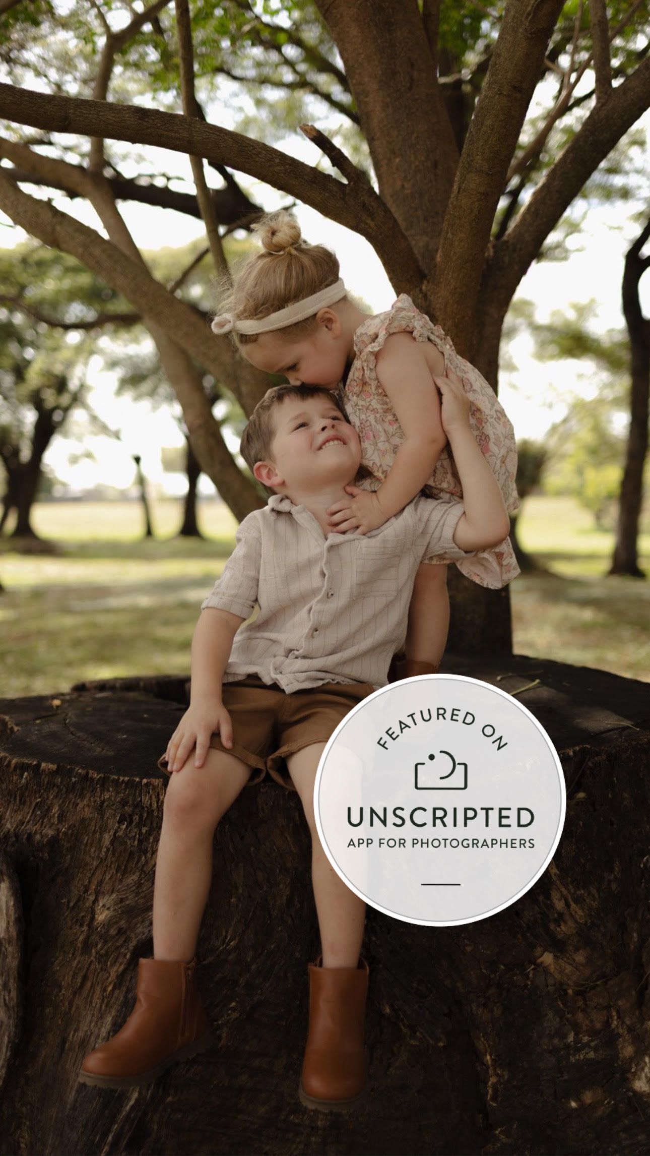 Children share a sweet moment while sitting on a large tree trunk in a sunlit park.