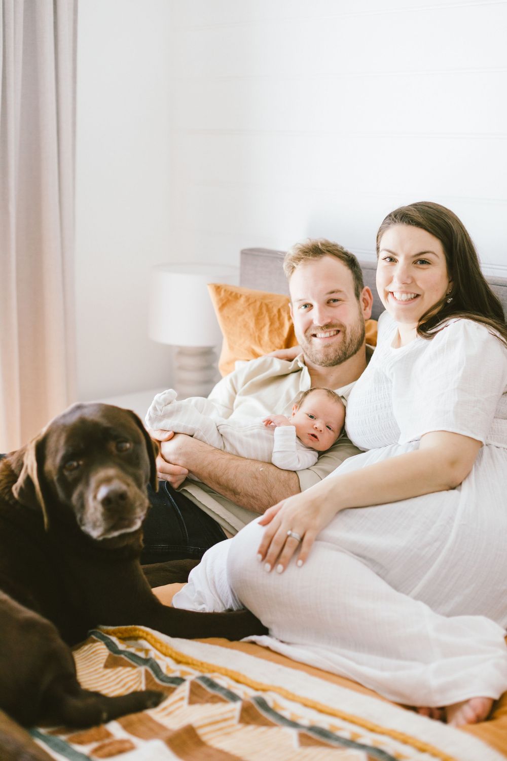Family lounges together on bed with their brown dog while holding sleeping newborn baby.