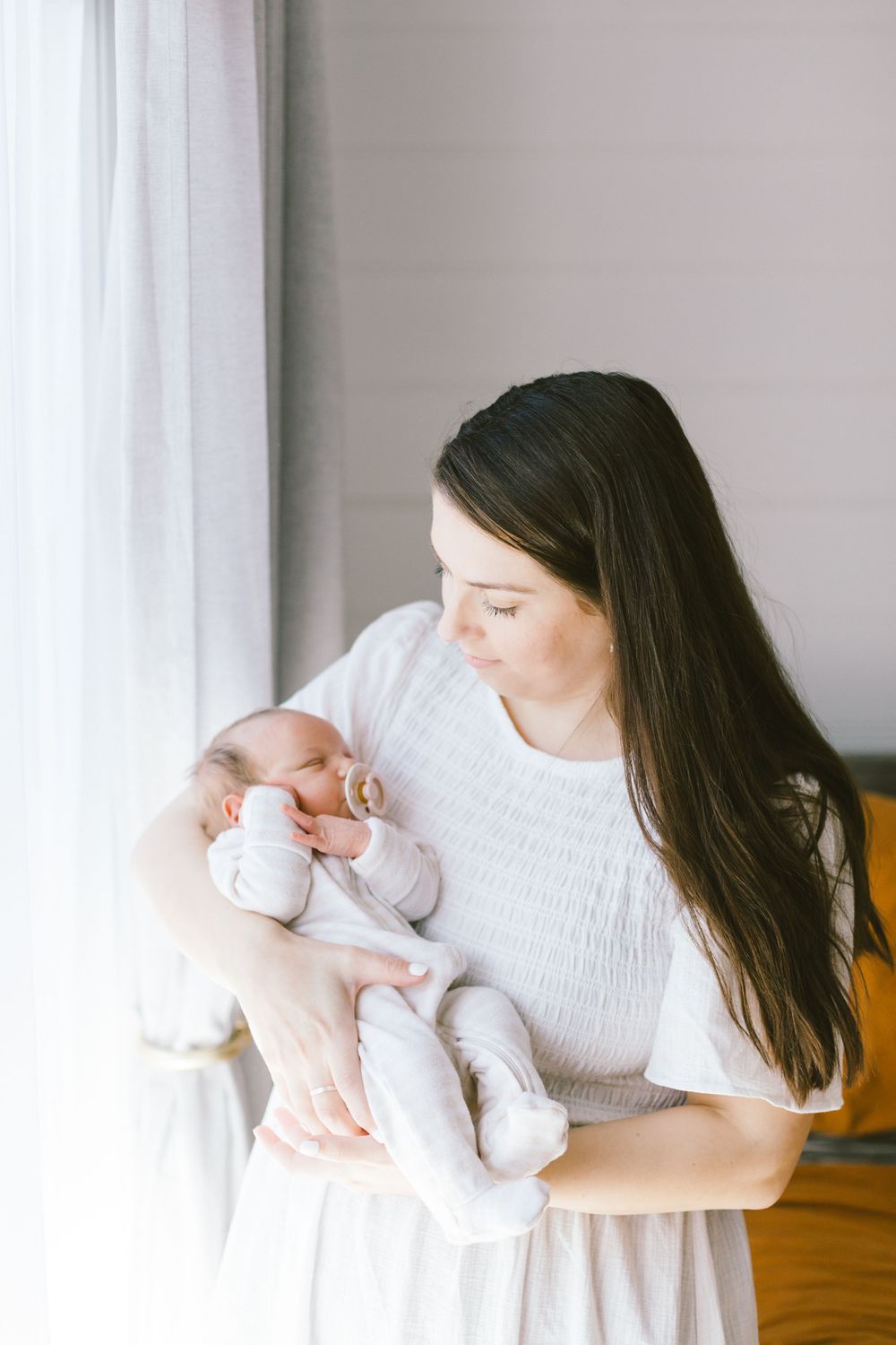 Person in white dress cradles sleeping newborn baby near window with natural light.