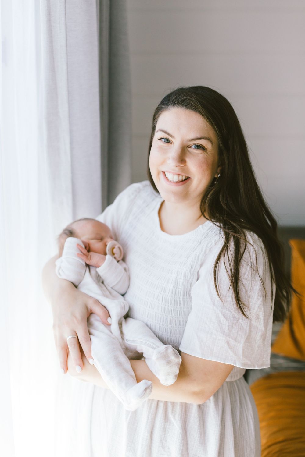 Person in white dress smiles while holding sleeping newborn baby near bright window.