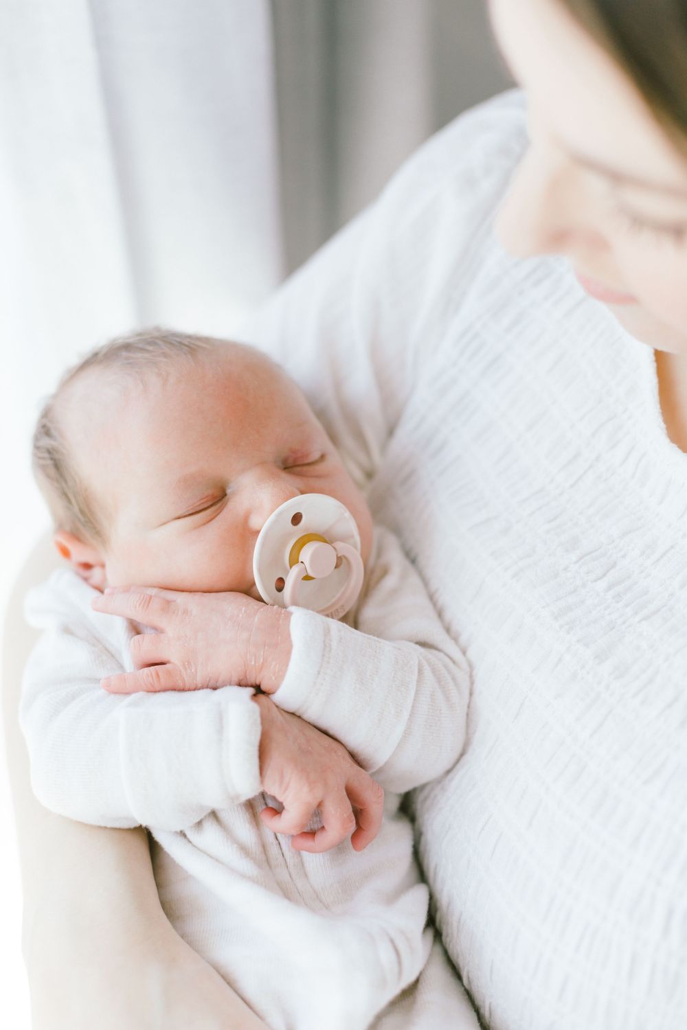 Close up of peaceful sleeping baby with pacifier resting against caregiver in white.