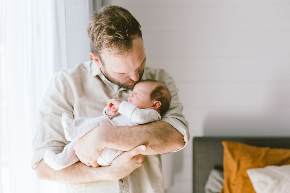 Parent in beige shirt gently cradles and gazes at newborn baby against white wall.
