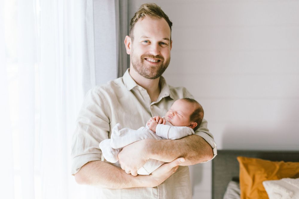 Parent smiles while holding sleeping newborn baby in casual home setting.