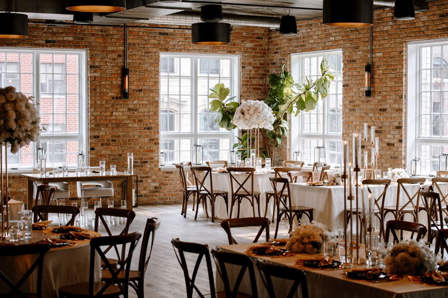 A rustic wedding reception venue with exposed brick walls, wooden cross-back chairs, and elegant table settings.