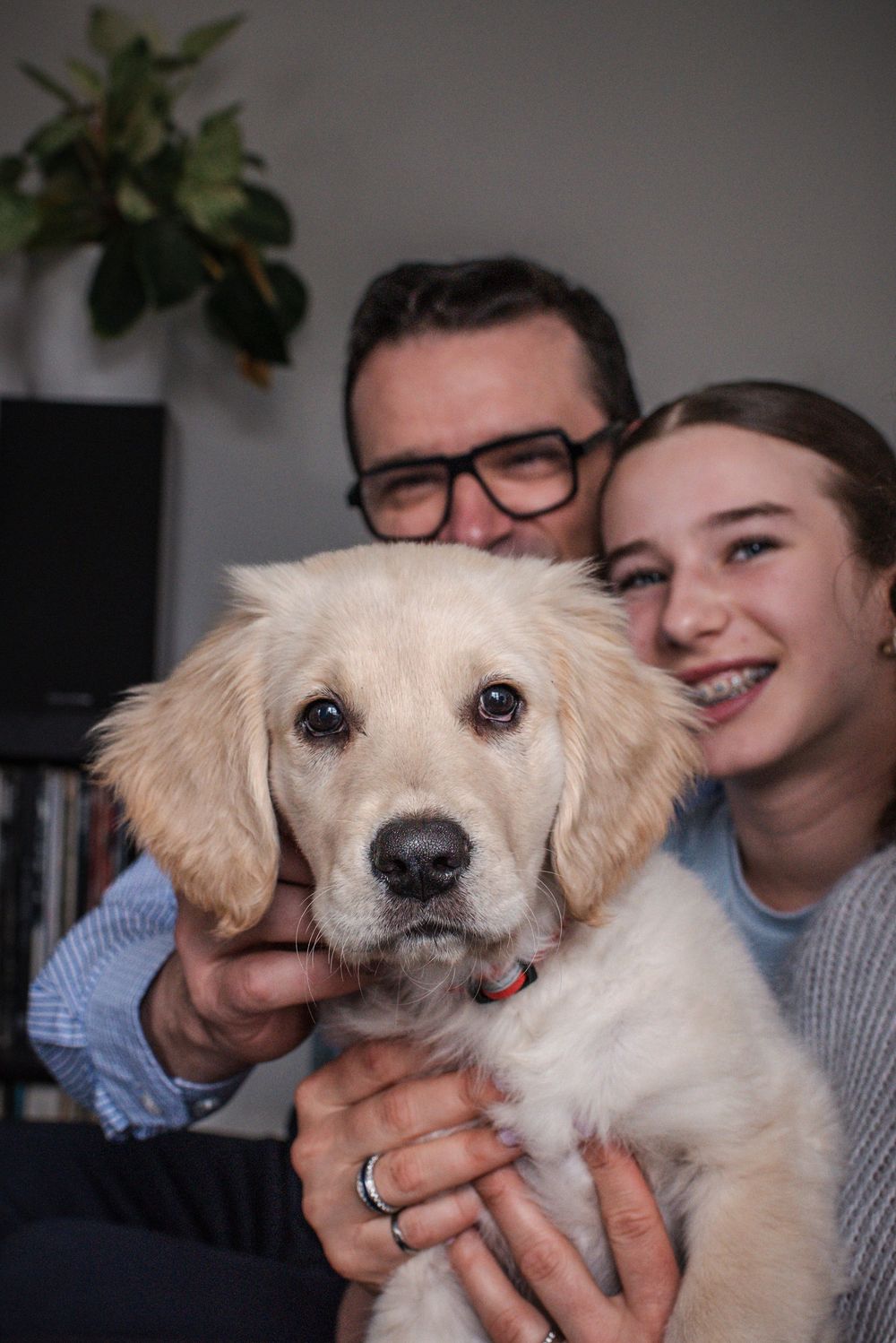 Father and daughter sit and smile at the camera from the couch as they hold on to their new puppy.