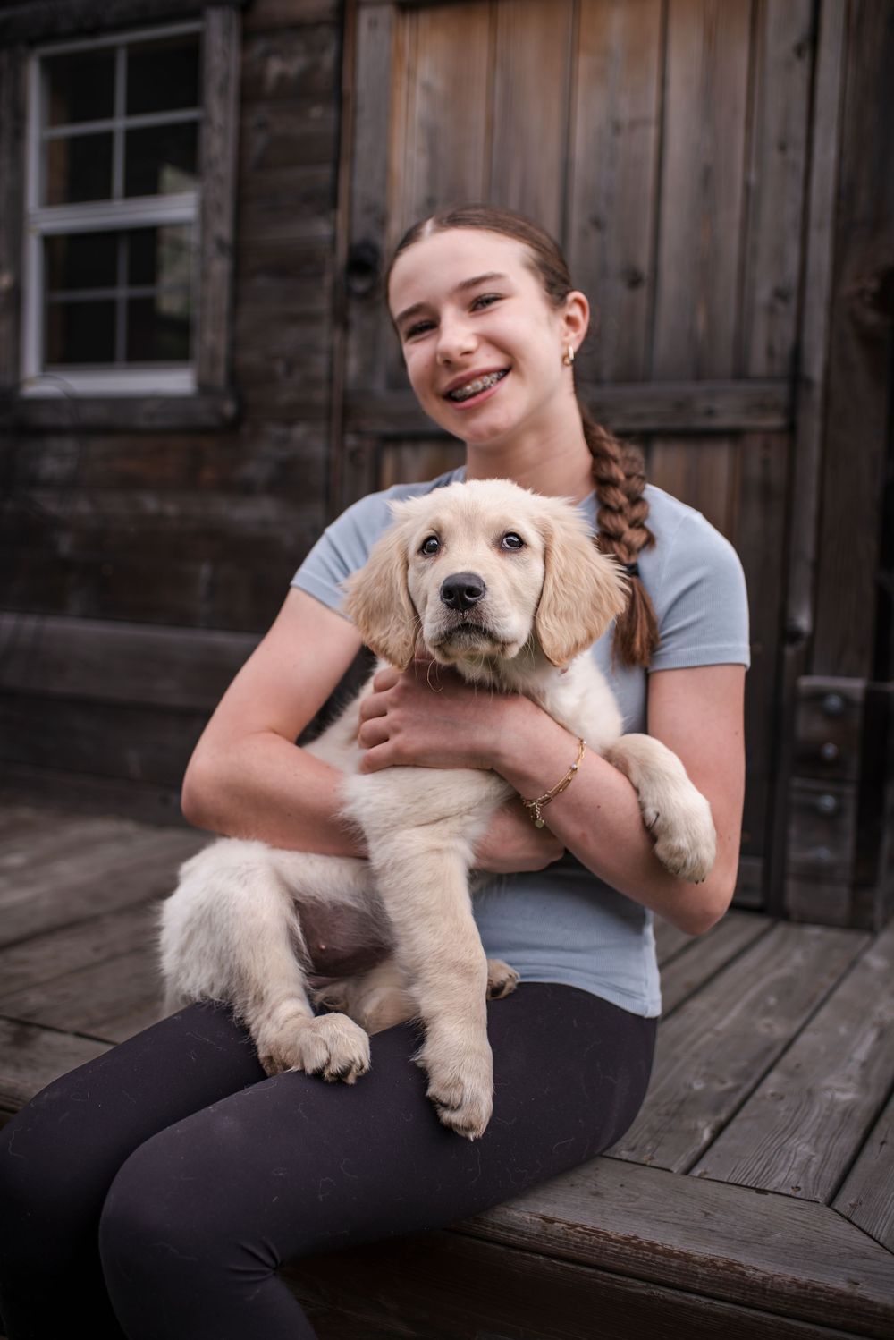 A girl holds her new puppy and smiles at the camera as she sits in front of her playhouse.