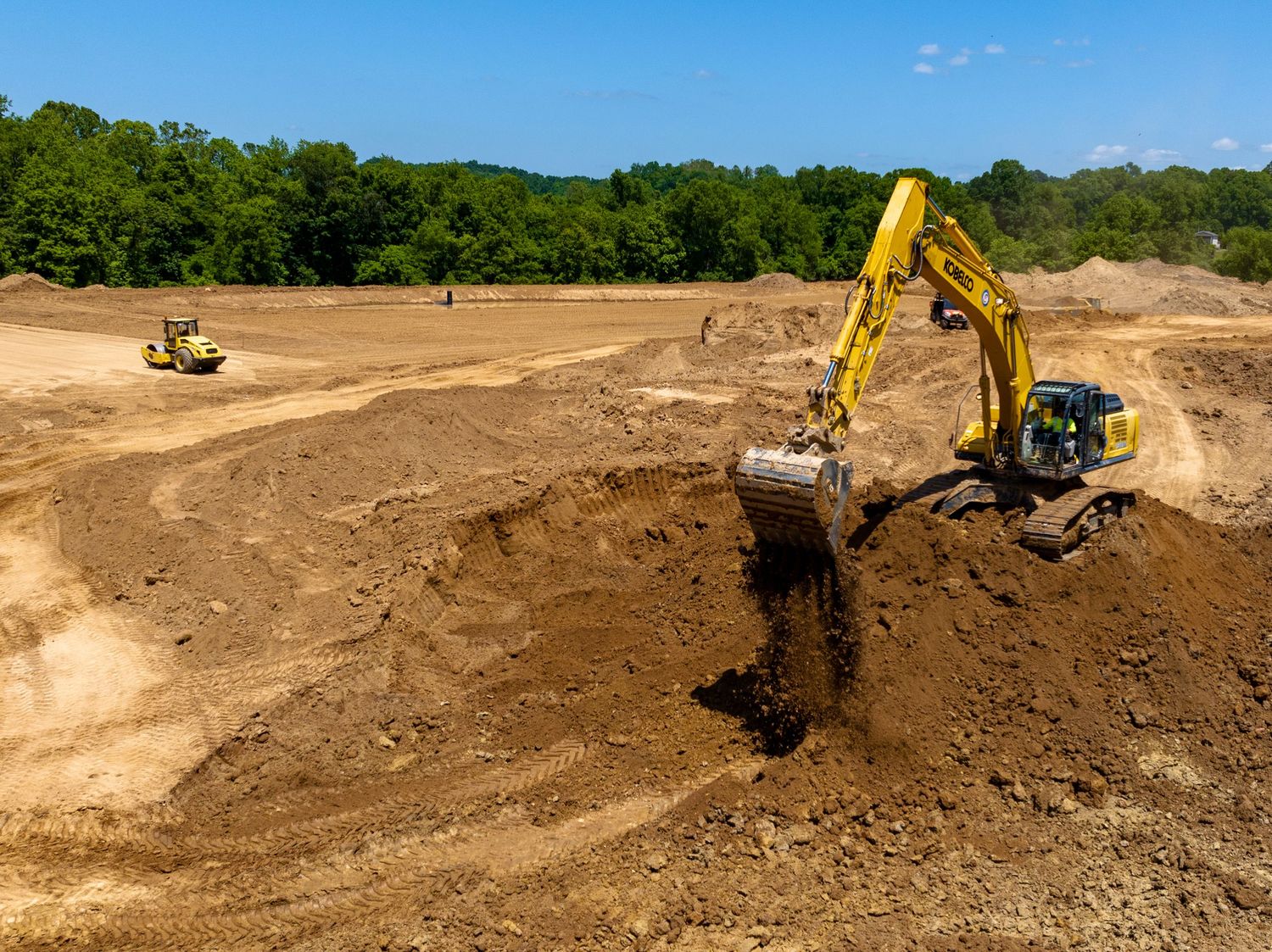 Yellow excavator digging dirt at construction site with earthmoving equipment on sunny day in wooded area.