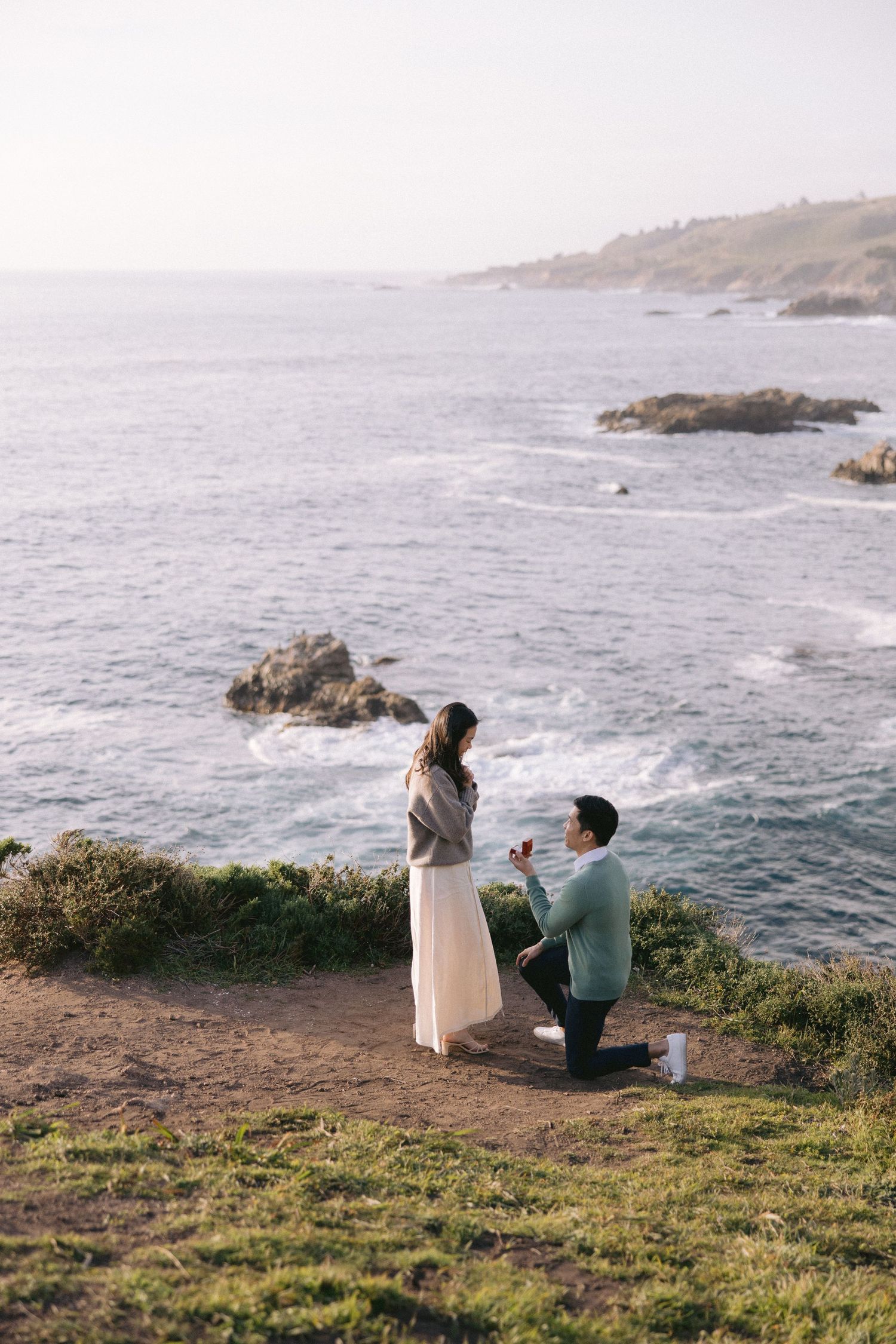 A romantic proposal scene on a coastal cliff overlooking the ocean with crashing waves and rocky outcrops at sunset.