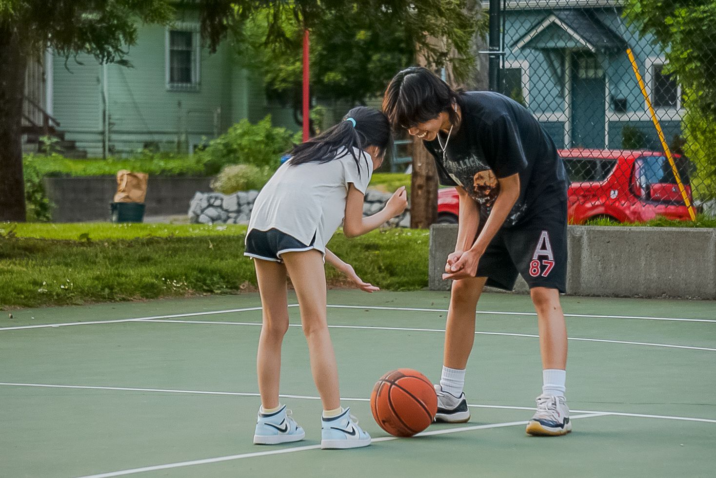 Two people play basketball together on an outdoor court on a sunny day, with one teaching dribbling techniques to the other.