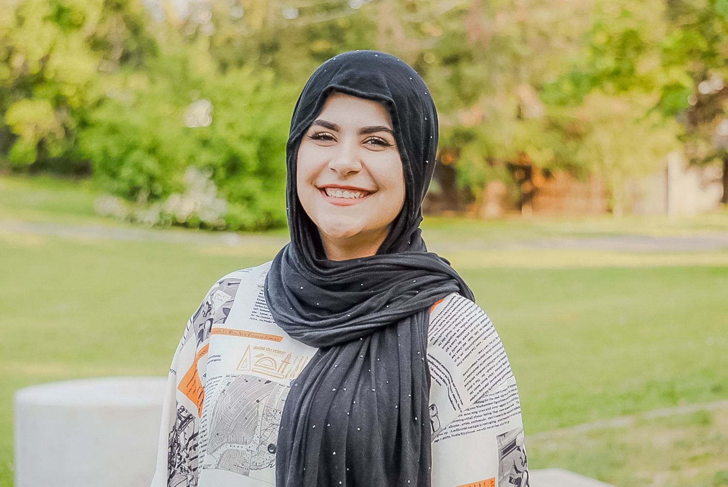 A cheerful person wearing a black hijab and patterned top smiles while standing in a sunny outdoor garden setting.