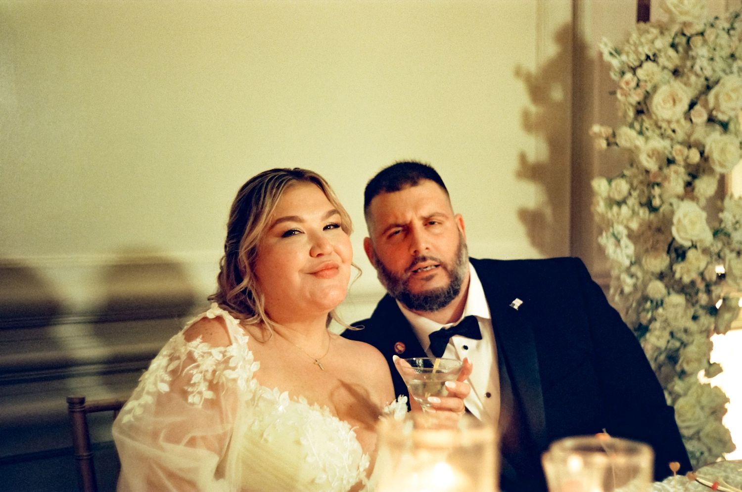 Romantic wedding photo shot on film showing newlywed couple sitting together in soft warm lighting with floral decor.