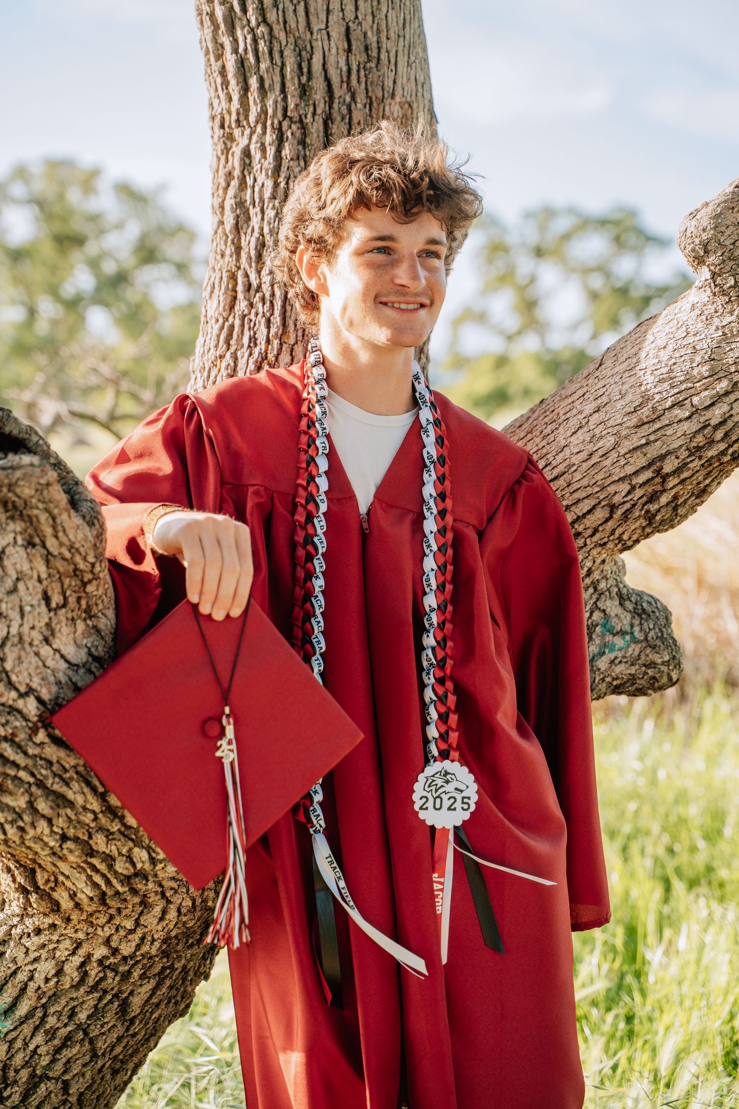 A graduate in red regalia smiles while posing with their diploma cap against a tree.