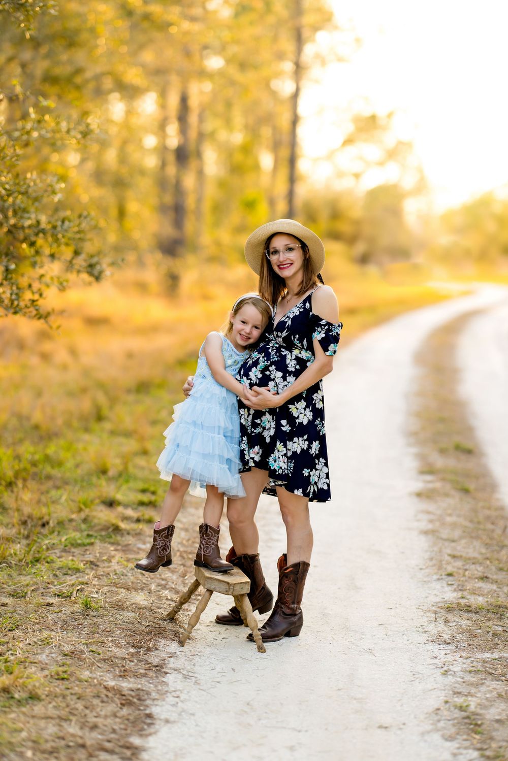 Two people in dresses and cowboy boots pose together on a sunlit dirt path.