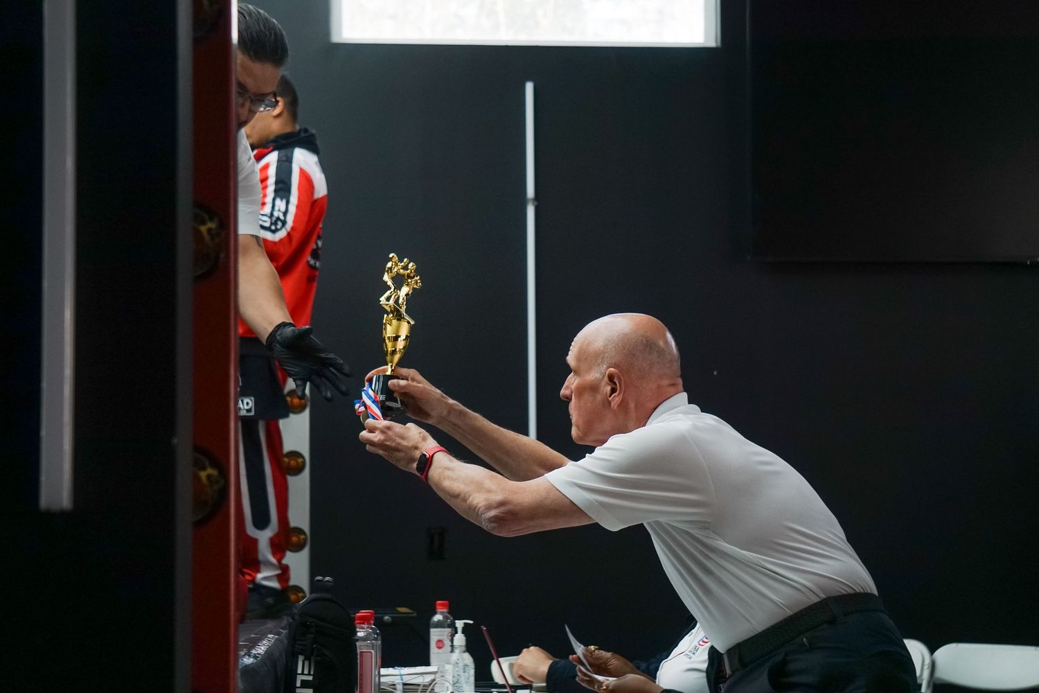 A person holds up a golden trophy while stretching towards a climbing wall in an indoor gym setting.