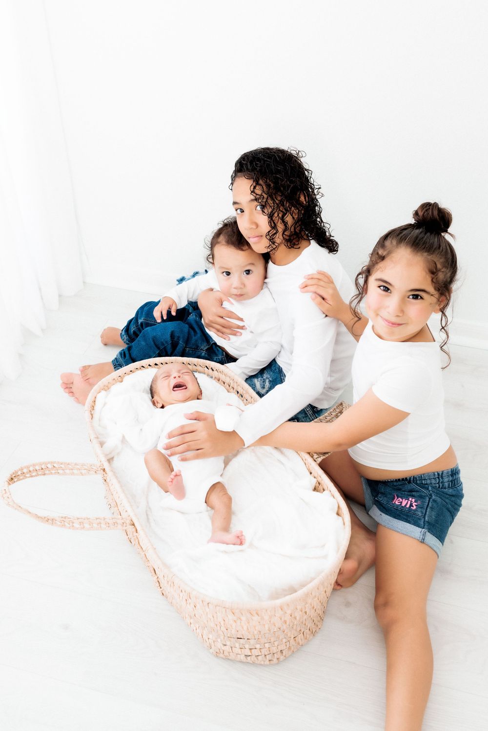 Mother sitting with two children and infant in wicker bassinet on white background.