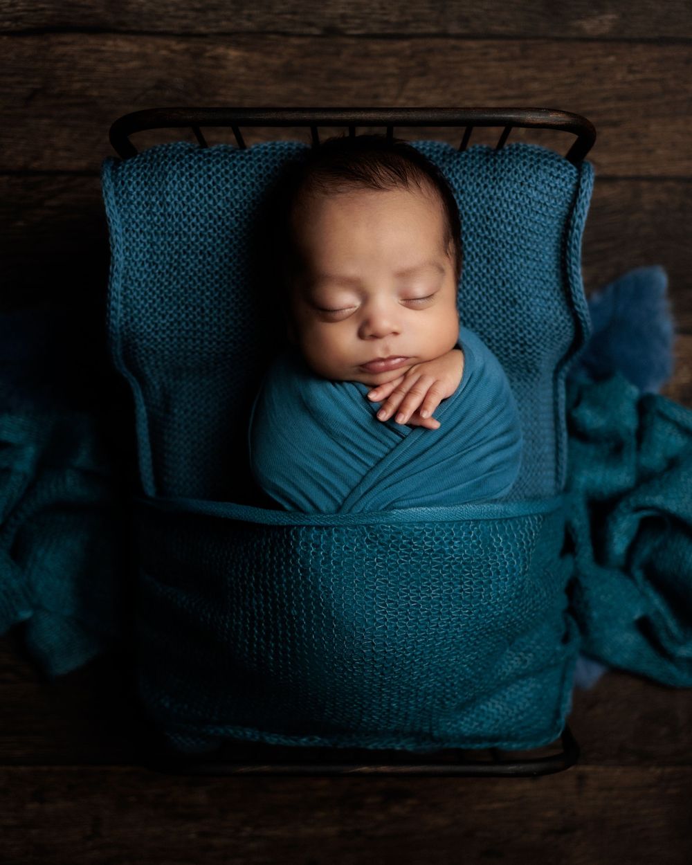 Sleeping newborn wrapped in teal swaddling blanket nestled in wooden basket with knitted teal cushioning.