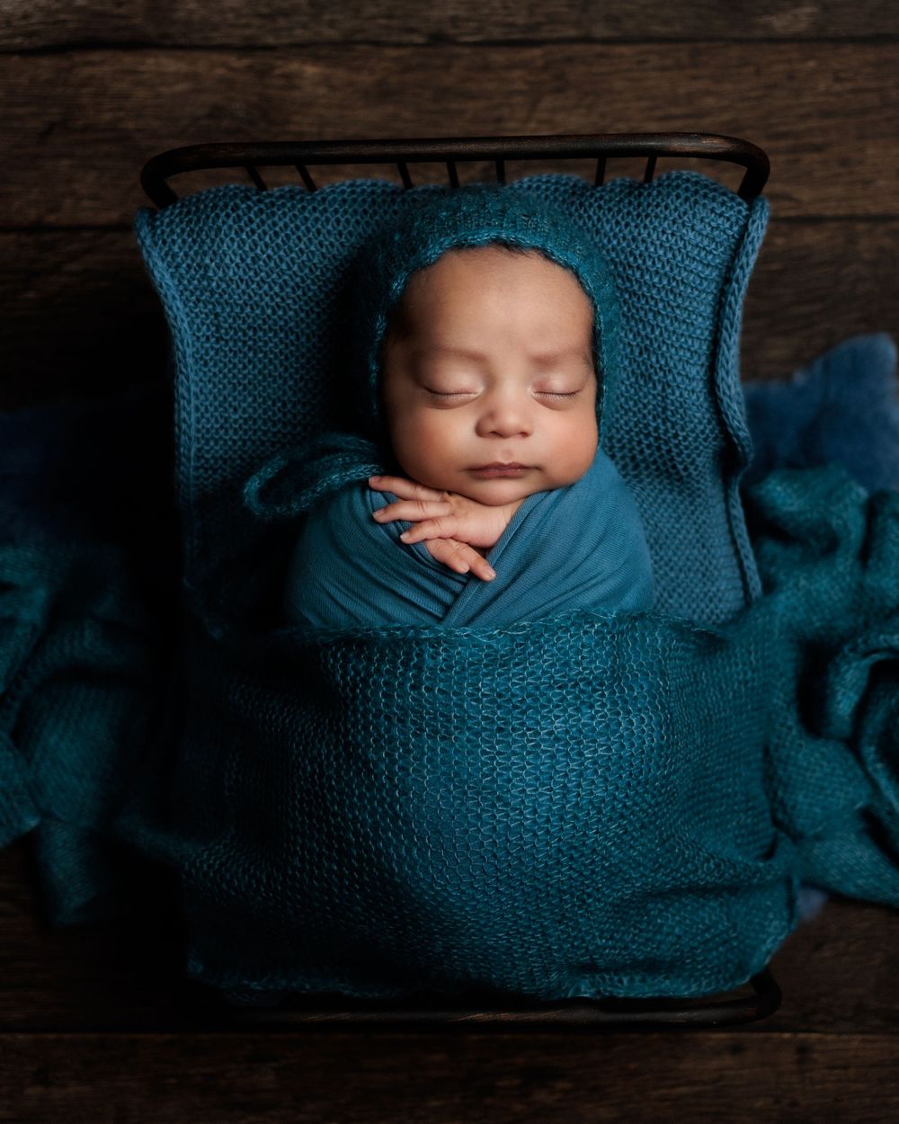 Sleeping newborn wrapped in soft teal knit blanket lying in wooden basket with matching textured fabric.