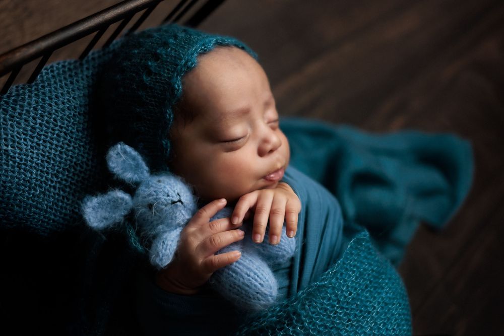 Sleeping baby in teal wrap cuddling small knitted blue bunny toy against dark wooden background.