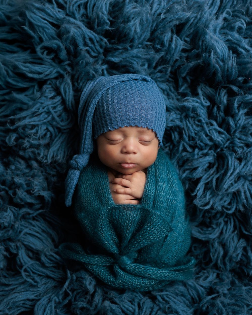 Sleeping newborn wrapped in cozy teal knit blanket wearing matching hat on plush blue faux fur backdrop.
