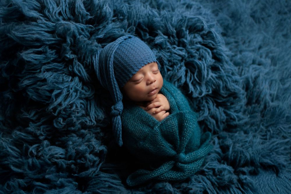 Peaceful sleeping newborn wrapped in cozy teal sweater and hat against matching textured backdrop.