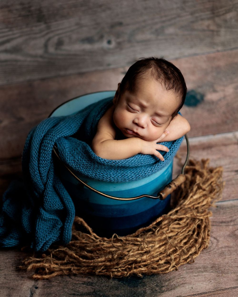 Sleeping newborn wrapped in teal knitted blanket rests peacefully in a bowl nestled on rustic wooden floor.