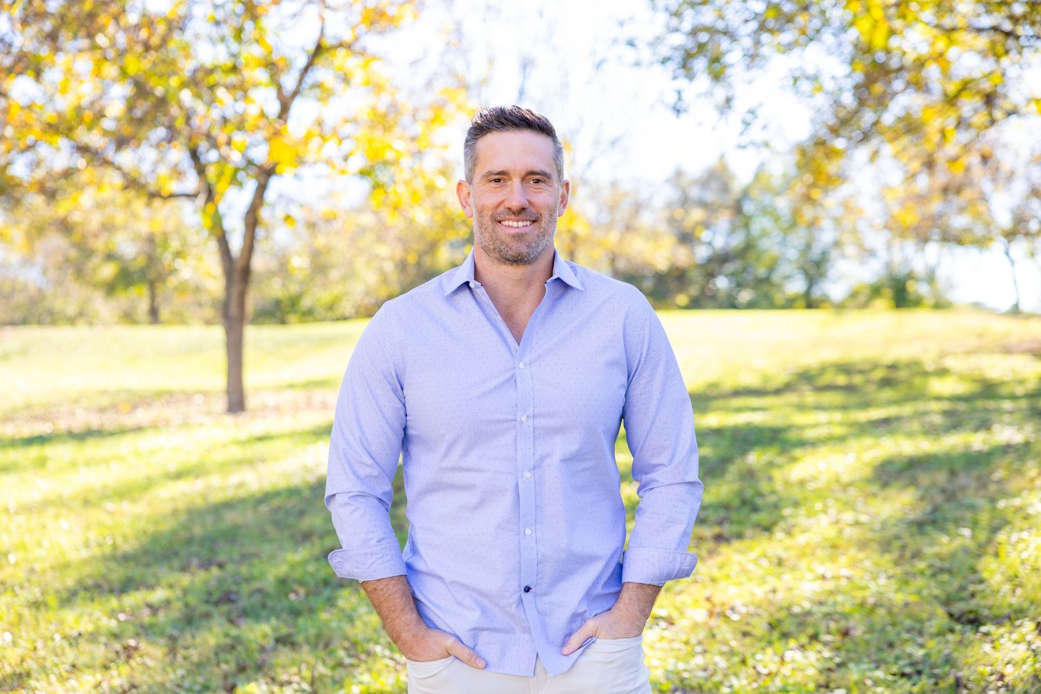 Someone wearing a light blue button-down shirt stands outdoors in a sunny park setting with autumn trees in background.
