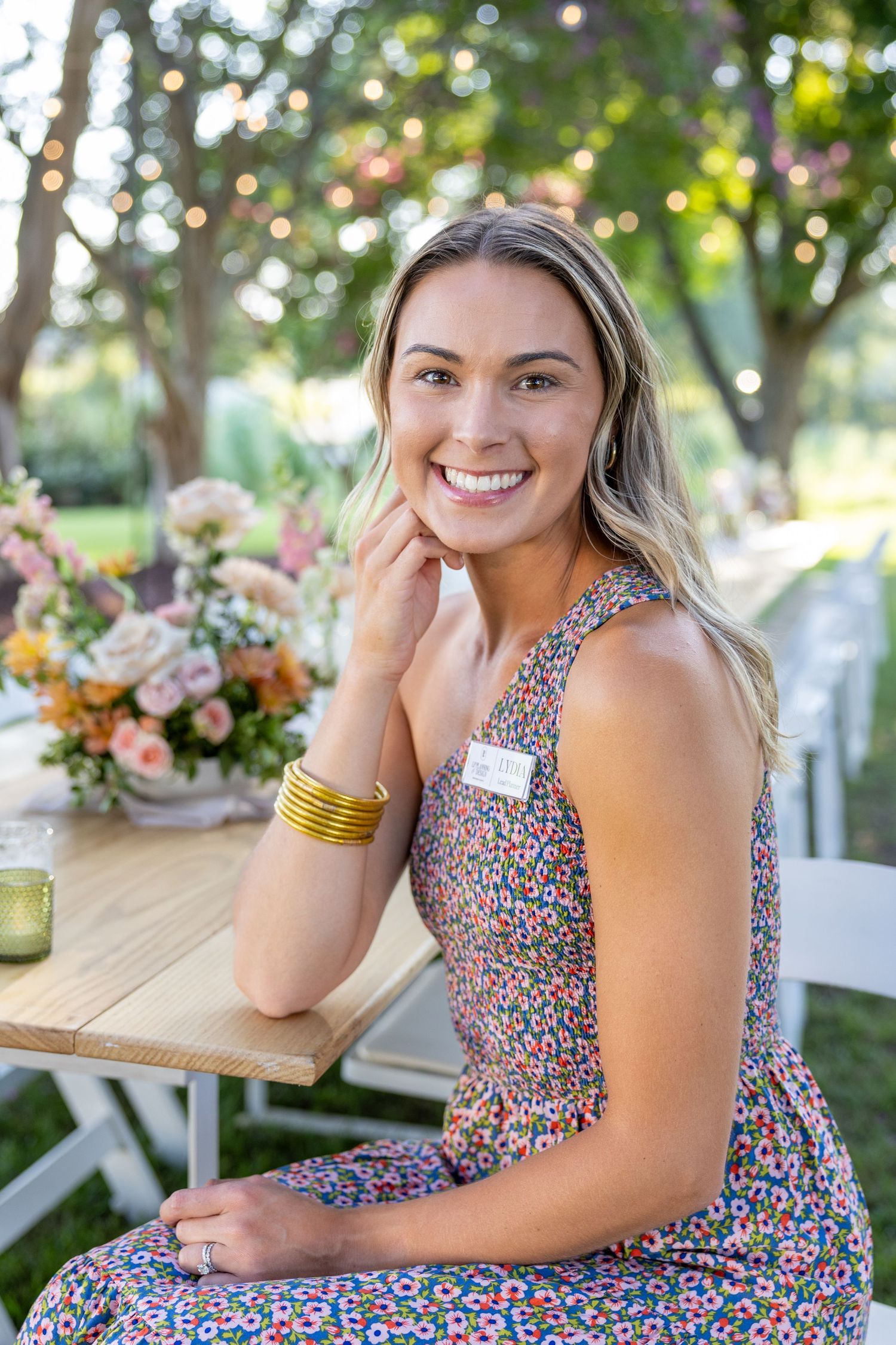 Woman in floral print dress sits at outdoor table with flower arrangements and golden bracelets.