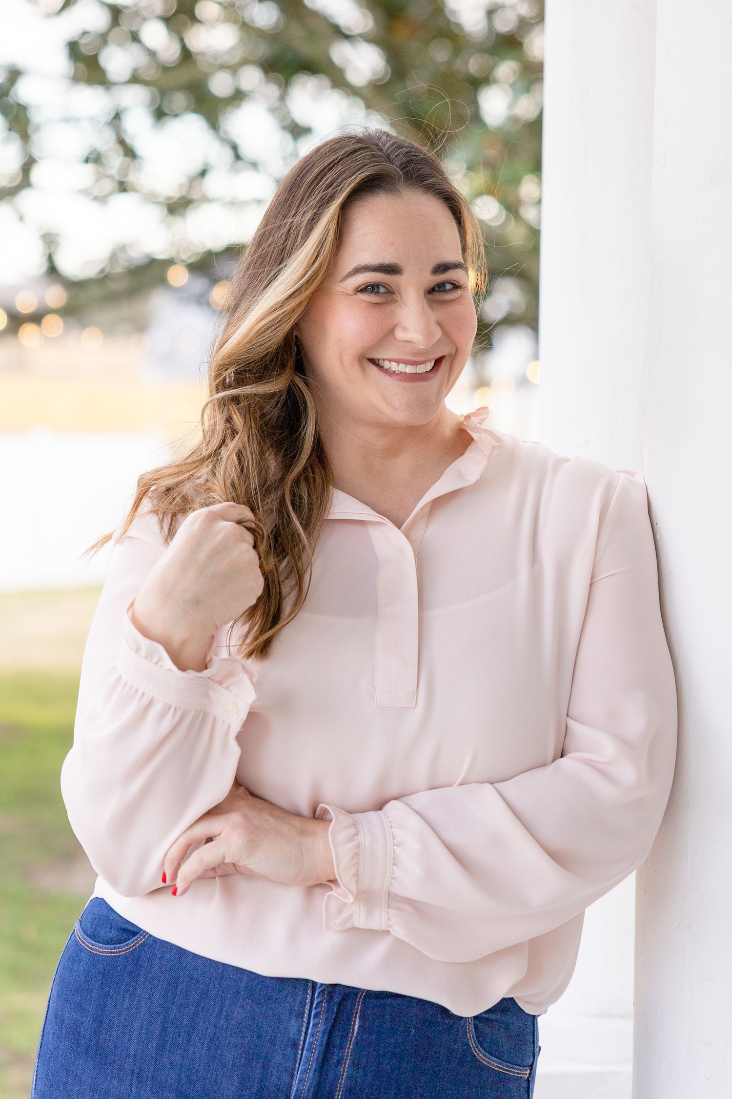 Woman in light pink pullover sweater and jeans smiles while standing outdoors near white column in her Clayton NC Headshot photos.