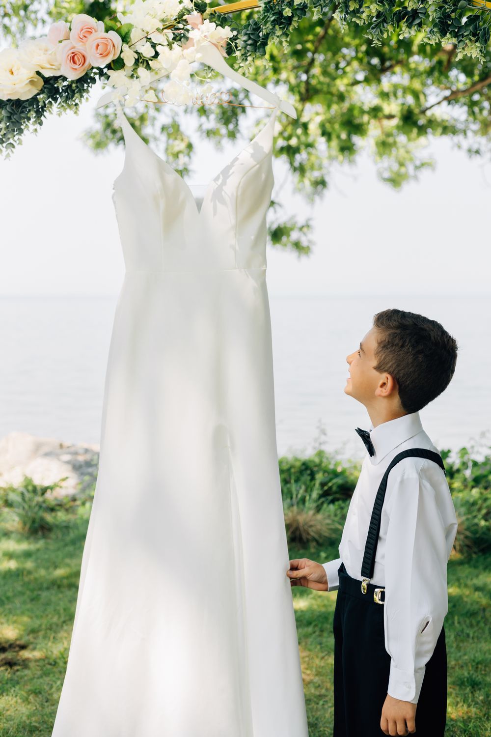A young ring bearer looking up at a white wedding dress hanging outdoors.