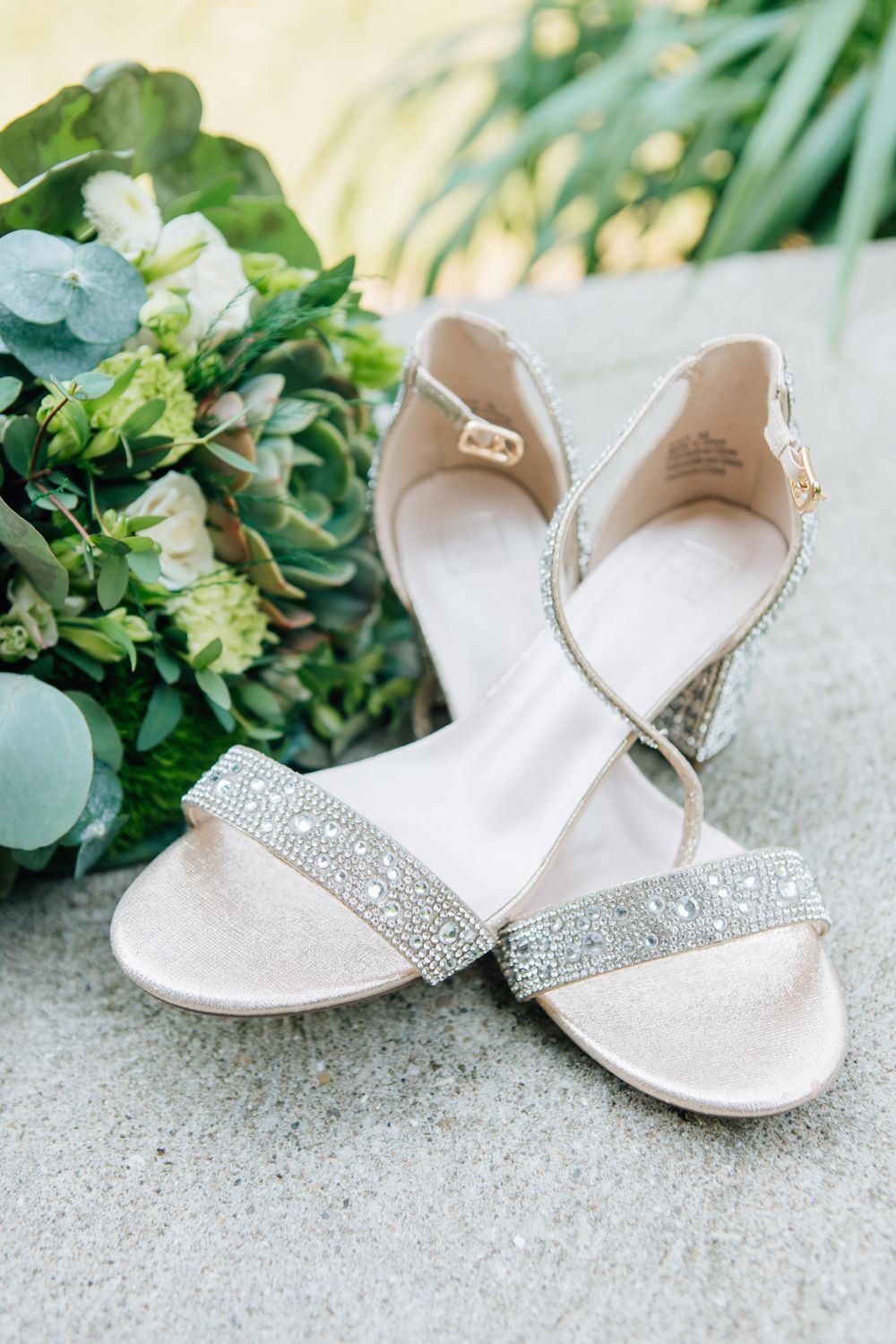 Elegant white bridal sandals with crystal embellishments displayed against a leafy green background.