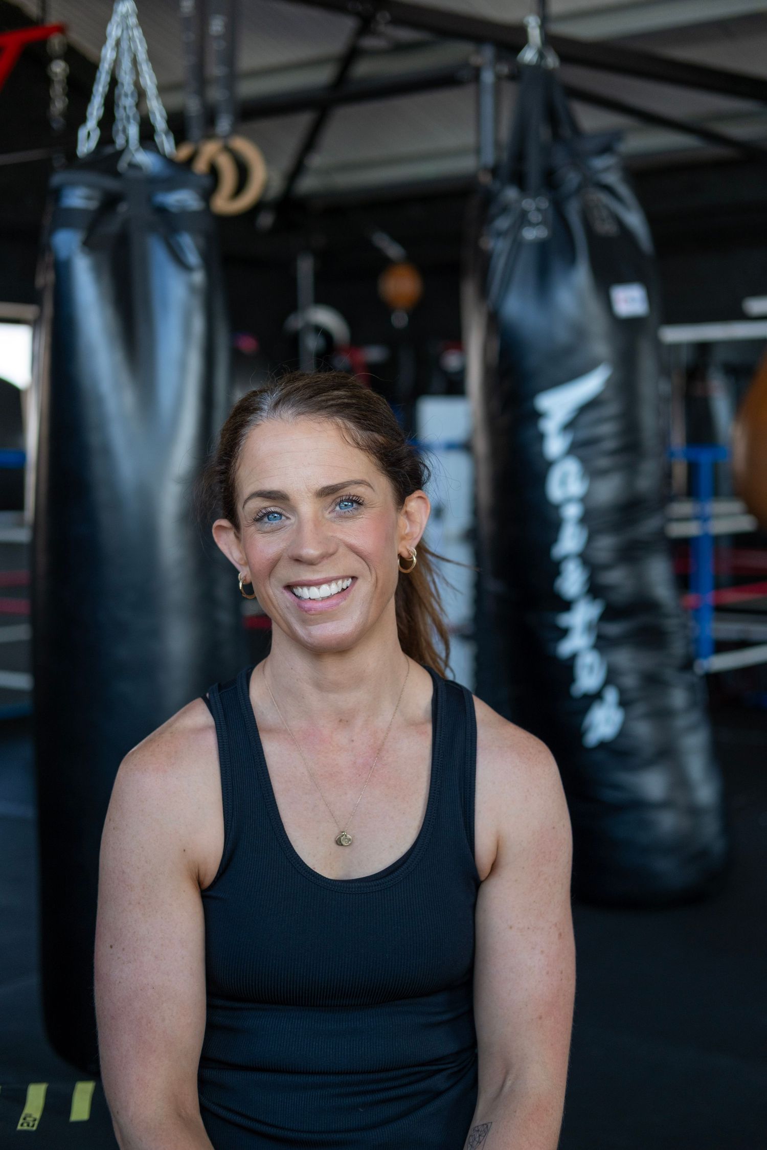 Fitness instructor poses in front of boxing bags at gym training area.