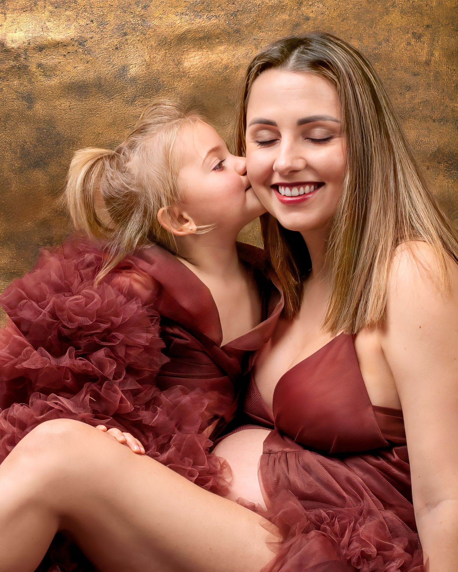 Motherhood portrait, mother and daughter in matching couture gowns, the girl kisses her mothers cheek both smiling.