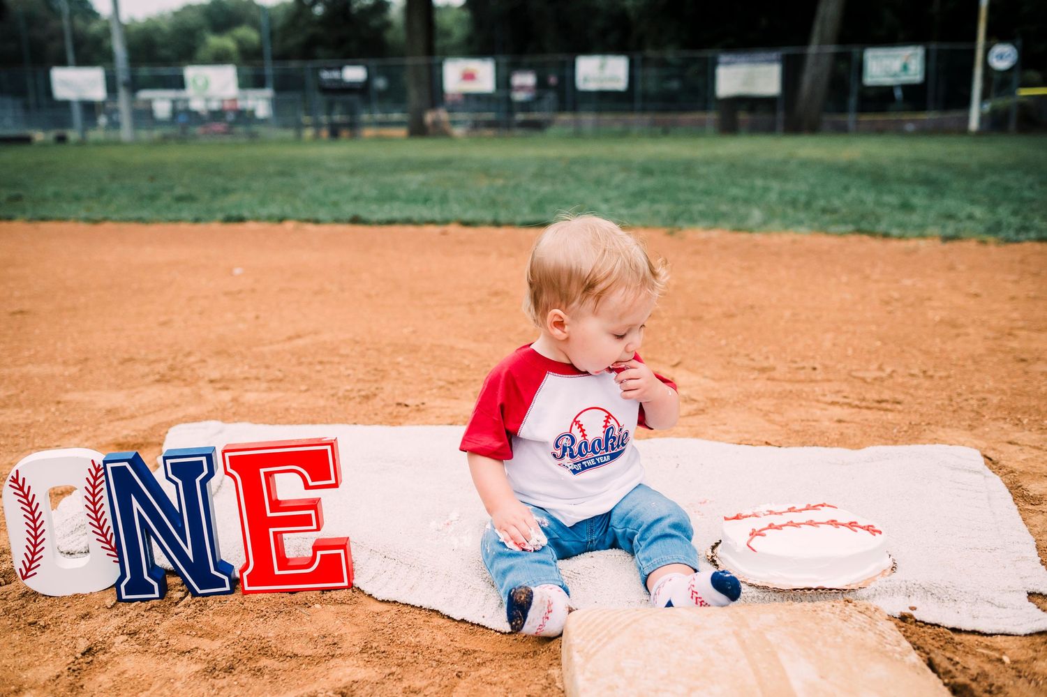 Small baby in baseball attire sits on white blanket at baseball field with red and blue lettering.