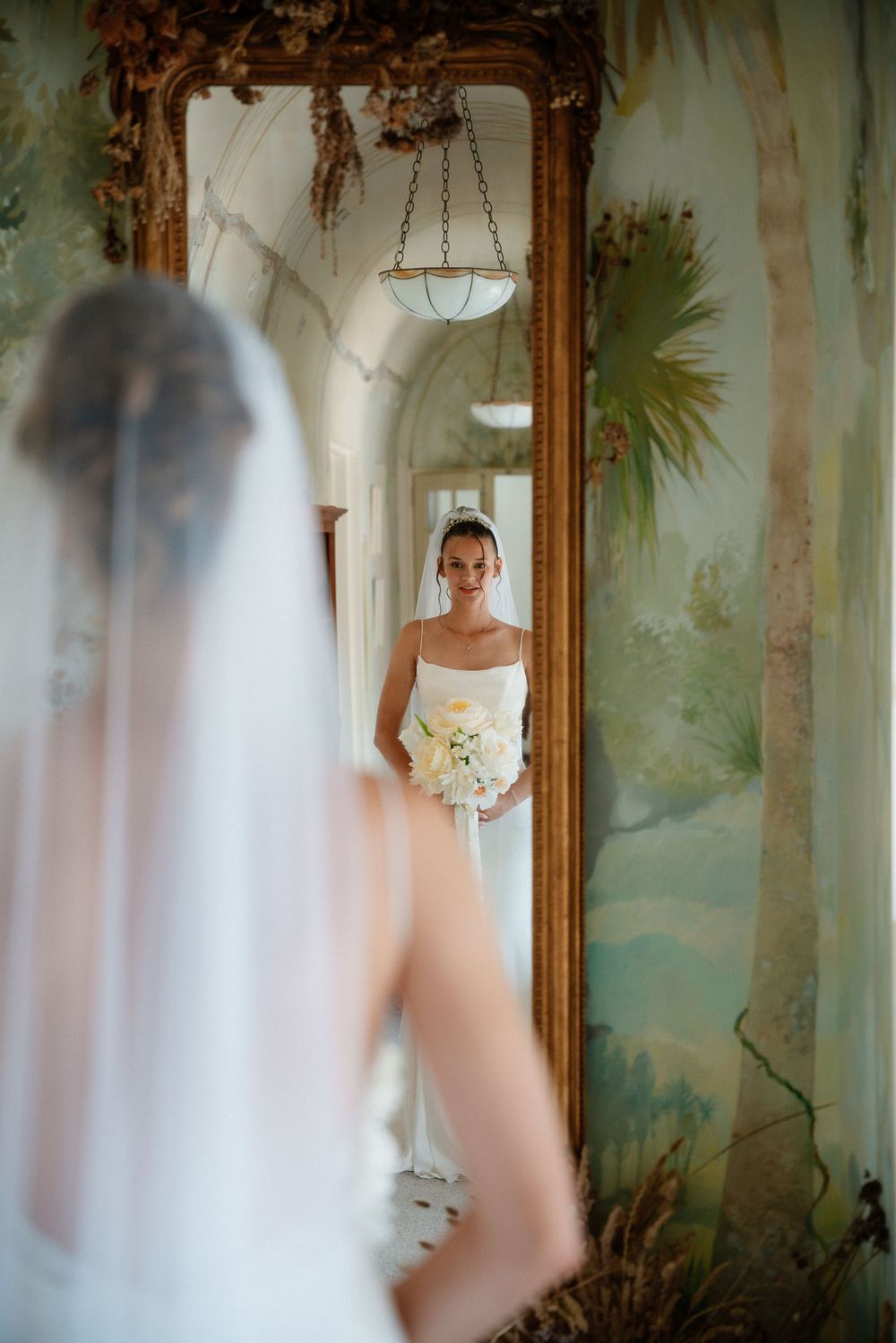 Bride in white dress holding bouquet reflected in ornate mirror with mural wall and vintage chandelier.