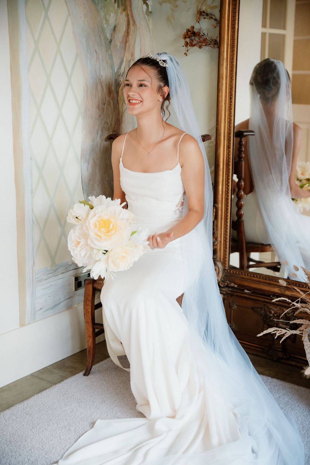Bride in elegant white spaghetti strap gown smiles while holding white bouquet next to antique mirror.