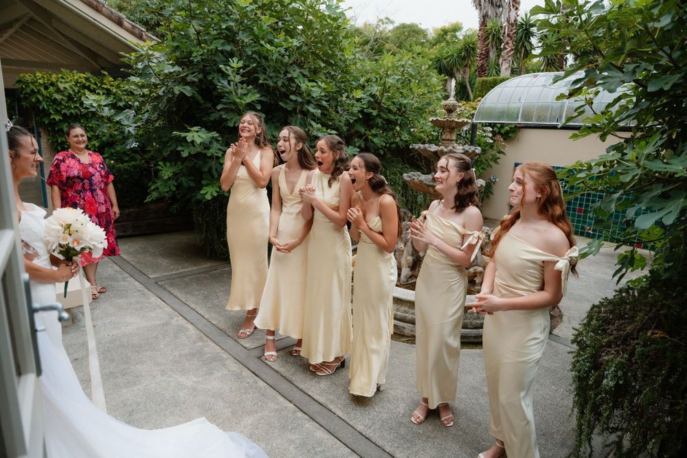 Bridesmaids in champagne colored dresses stand in line outdoors at wedding ceremony.
