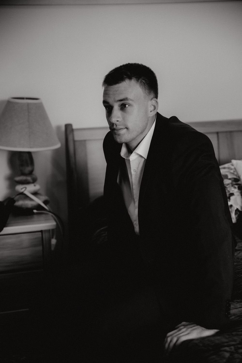 Elegant black and white portrait of a person in formal attire sitting pensively beside a bedroom lamp.