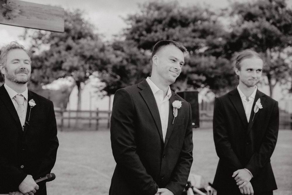 Black and white photo of three groomsmen in formal suits standing outdoors at a wedding ceremony.