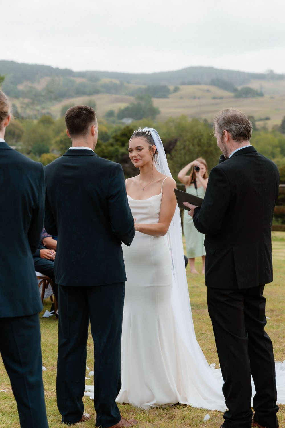 An outdoor wedding ceremony takes place on a hilltop with rolling countryside views in the background.