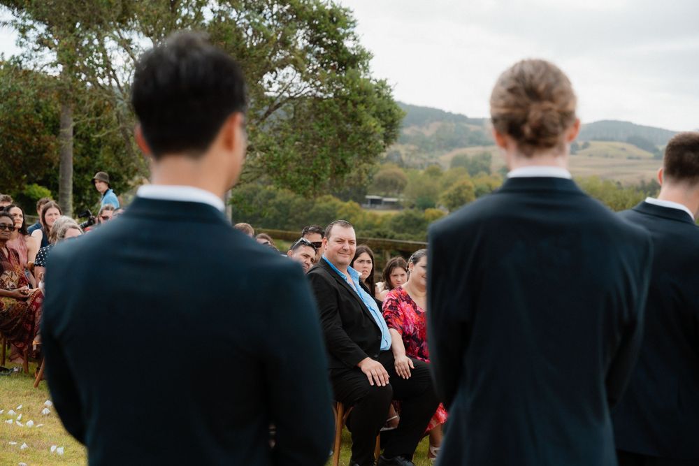 Wedding guests look on and smile during an outdoor ceremony with trees and hills in the background.